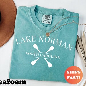 May include: Seafoam green t-shirt with the text "Lake Norman North Carolina" and crossed oars in white. A gold necklace with a pendant rests on the shirt. The shirt has a Comfort Colors tag.