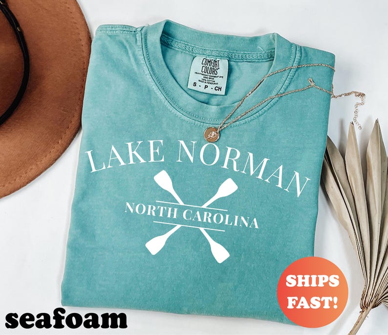 May include: Seafoam green t-shirt with the text "Lake Norman North Carolina" and crossed oars in white. A gold necklace with a pendant rests on the shirt. The shirt has a Comfort Colors tag.
