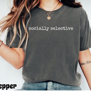 May include: A dark gray t-shirt with the words "socially selective" printed in white. The shirt is a crew neck and appears to be made of a soft material. The word "pepper" is printed in the bottom left corner.