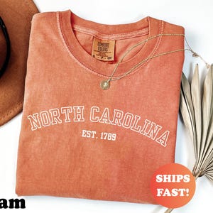 Puede incluir: Una camiseta de color coral con las palabras "NORTH CAROLINA" y "EST. 1789" en letras blancas. La camiseta tiene cuello redondo y una etiqueta peque&ntilde;a. Un collar dorado y un sombrero marr&oacute;n tambi&eacute;n est&aacute;n en la imagen.