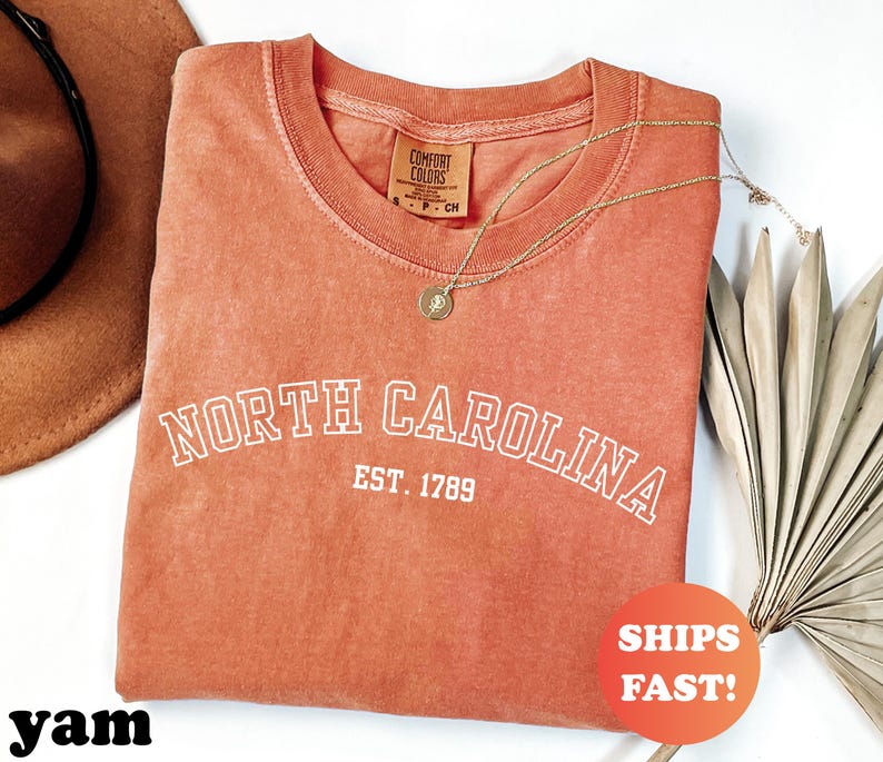 Puede incluir: Una camiseta de color coral con las palabras "NORTH CAROLINA" y "EST. 1789" en letras blancas. La camiseta tiene cuello redondo y una etiqueta peque&ntilde;a. Un collar dorado y un sombrero marr&oacute;n tambi&eacute;n est&aacute;n en la imagen.
