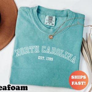Puede incluir: Camiseta verde mar con las palabras "NORTH CAROLINA" y "EST. 1789" impresas en blanco. Un collar dorado con un colgante descansa sobre la camiseta. La camiseta est&aacute; doblada y tiene una etiqueta Comfort Colors. Un c&iacute;rculo naranja dice "SHIPS FAST!"