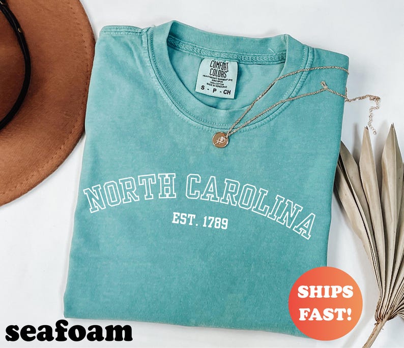 Puede incluir: Camiseta verde mar con las palabras "NORTH CAROLINA" y "EST. 1789" impresas en blanco. Un collar dorado con un colgante descansa sobre la camiseta. La camiseta est&aacute; doblada y tiene una etiqueta Comfort Colors. Un c&iacute;rculo naranja dice "SHIPS FAST!"