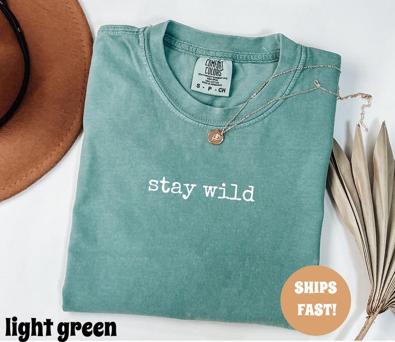 May include: A light green t-shirt with the words "stay wild" in white lettering. A gold necklace with a pendant rests on the shirt. The shirt is folded and has a tag that says "Comfort Colors".