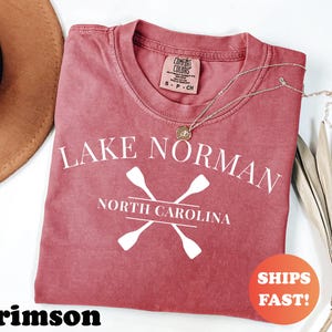 May include: Crimson-colored t-shirt with the text "Lake Norman North Carolina" and crossed oars in white. The shirt has a crew neck and a small tag at the neckline. A gold necklace is draped across the shirt.