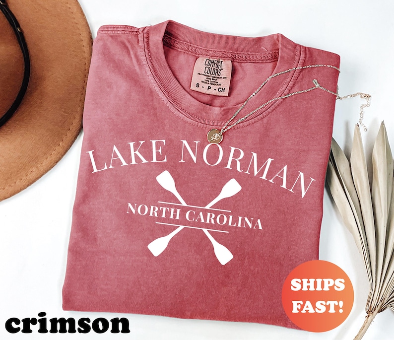 May include: Crimson-colored t-shirt with the text "Lake Norman North Carolina" and crossed oars in white. The shirt has a crew neck and a small tag at the neckline. A gold necklace is draped across the shirt.