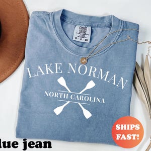 May include: A blue jean colored t-shirt with the words "Lake Norman North Carolina" in white, along with crossed oars. A gold necklace and a brown hat are also in the image. The shirt has a Comfort Colors tag.