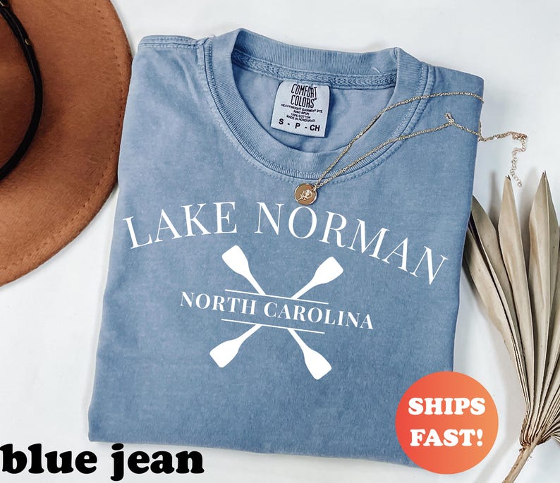 May include: A blue jean colored t-shirt with the words "Lake Norman North Carolina" in white, along with crossed oars. A gold necklace and a brown hat are also in the image. The shirt has a Comfort Colors tag.
