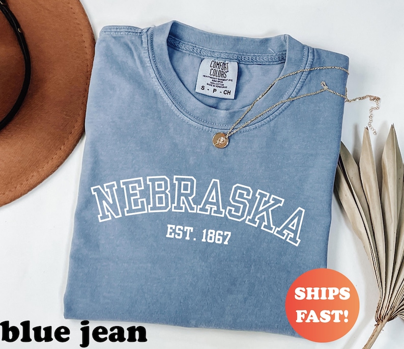 May include: A blue jean colored t-shirt with the word "NEBRASKA" in white block letters and "EST. 1867" underneath. A gold necklace with a pendant is draped across the shirt. A brown hat and dried plant are in the background.