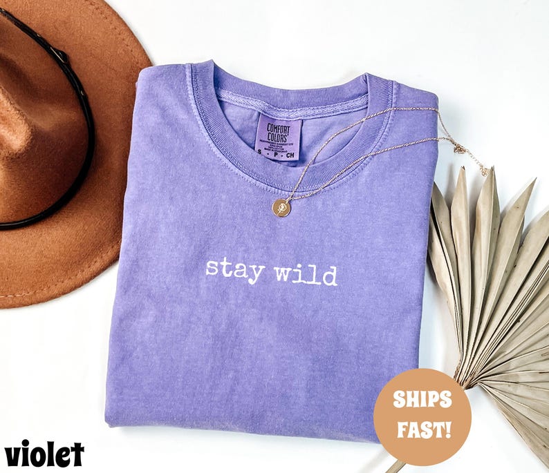 May include: A violet Comfort Colors t-shirt with the phrase "stay wild" in white script. A gold necklace with a pendant is draped over the shirt. A brown hat is in the background.