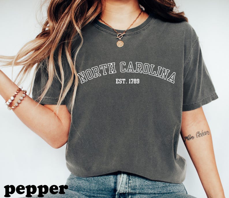Puede incluir: Camiseta gris oscuro con el texto blanco "NORTH CAROLINA EST. 1789" en arco. La camiseta es de estilo casual. La palabra "pepper" es visible en la parte inferior de la imagen.