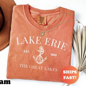 May include: A coral-colored t-shirt with the text "LAKE ERIE" and an anchor graphic. The shirt also features the text "EST. 1669" and "THE GREAT LAKES". A gold necklace and a brown hat are visible. A sticker that says "SHIPS FAST!" is also visible.