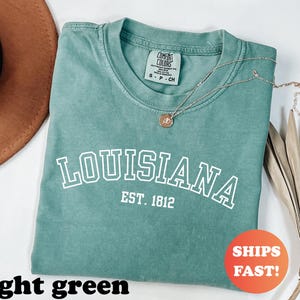 May include: A light green t-shirt with the word "LOUISIANA" in white block letters, with "EST. 1812" underneath. A gold necklace with a pendant rests on the shirt. The shirt is folded, and a brown hat is visible.