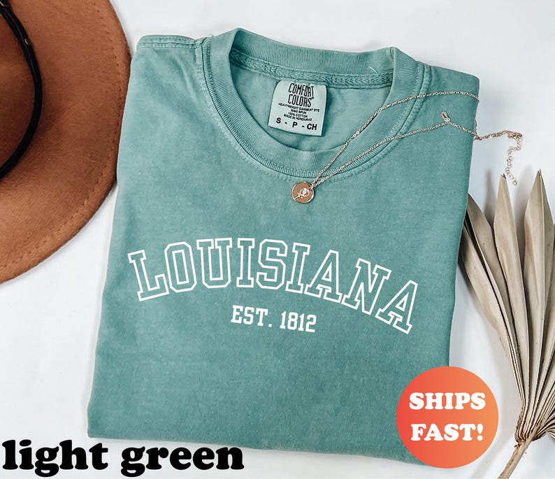 May include: A light green t-shirt with the word "LOUISIANA" in white block letters, with "EST. 1812" underneath. A gold necklace with a pendant rests on the shirt. The shirt is folded, and a brown hat is visible.