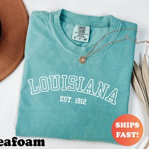 May include: Seafoam green t-shirt with the word "LOUISIANA" in white block letters and "EST. 1812" underneath. A gold necklace with a pendant rests on the shirt. The shirt is folded and has a "SHIPS FAST!" sticker.