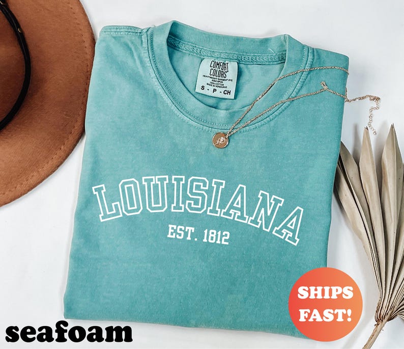 May include: Seafoam green t-shirt with the word "LOUISIANA" in white block letters and "EST. 1812" underneath. A gold necklace with a pendant rests on the shirt. The shirt is folded and has a "SHIPS FAST!" sticker.