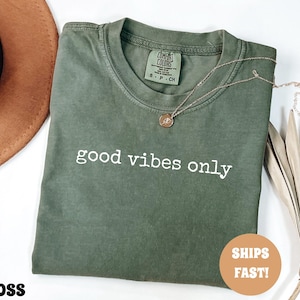 May include: A moss green t-shirt with the words "good vibes only" printed in white. A gold necklace with a pendant is on the shirt. The shirt is folded and displayed with a brown hat and dried plant.