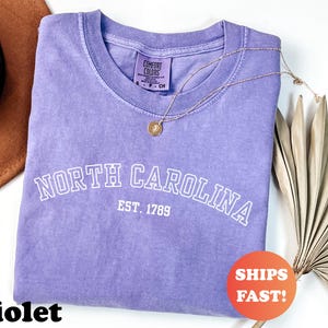 Puede incluir: Una camiseta violeta Comfort Colors con la inscripci&oacute;n blanca "NORTH CAROLINA EST. 1789". Un collar dorado con un colgante descansa sobre la camiseta. Una pegatina naranja con "SHIPS FAST!" es visible.