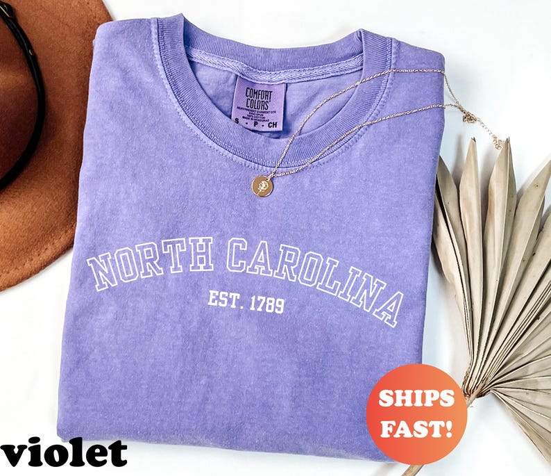 Puede incluir: Una camiseta violeta Comfort Colors con la inscripci&oacute;n blanca "NORTH CAROLINA EST. 1789". Un collar dorado con un colgante descansa sobre la camiseta. Una pegatina naranja con "SHIPS FAST!" es visible.