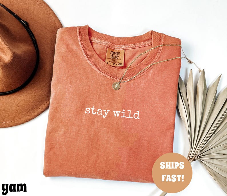 May include: A coral-colored t-shirt with the words "stay wild" in white script. A gold necklace with a pendant rests on the shirt. A brown cowboy hat and dried palm leaves are in the background.