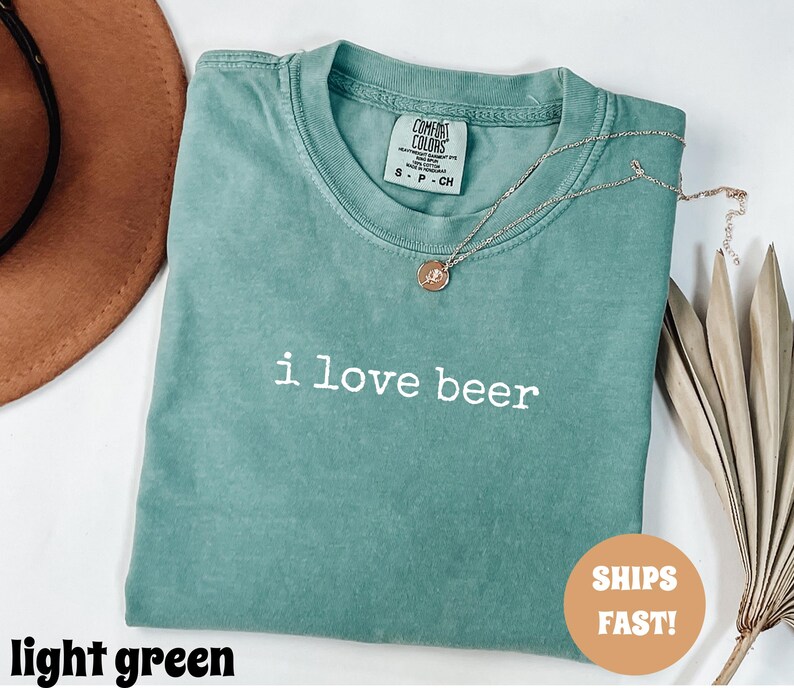 May include: A light green t-shirt with the text "i love beer" in white. A gold necklace with a pendant rests on the shirt. The shirt is folded and has a tag that says "COMFORT COLORS".