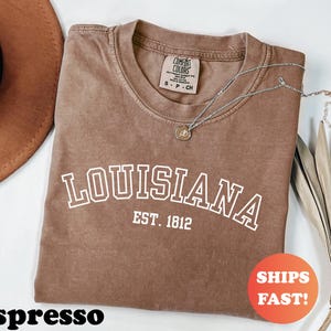 May include: Espresso-colored t-shirt with the word "LOUISIANA" in white block letters and "EST. 1812" underneath. A silver necklace with a pendant rests on the shirt. The shirt has a Comfort Colors tag.