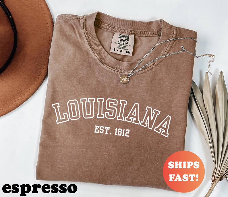 May include: Espresso-colored t-shirt with the word "LOUISIANA" in white block letters and "EST. 1812" underneath. A silver necklace with a pendant rests on the shirt. The shirt has a Comfort Colors tag.