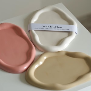 Könnte beinhalten: Drei ovale Tabletts in Rosa, Weiß und Beige. Das weiße Tablett hat ein Papierband mit der Aufschrift "Oval Cloud Tray" und "Eco Friendly, VOC Free, Durable, Non-Toxic". Die Tabletts haben ein wellenförmiges, wolkenartiges Design.