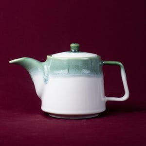 以下が含まれることがあります： 白ベースに緑釉の上部と取っ手が付いた陶器のティーポット。蓋には小さな緑色のつまみがあります。ティーポットは、注ぎやすいように注ぎ口と取っ手がデザインされています。背景は無地のマルーン色です。