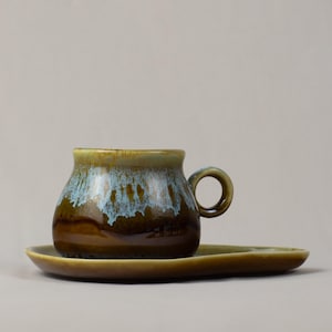 Puede incluir: Un juego de taza y platillo de café de cerámica. La taza tiene forma redondeada con asa, y el platillo es ovalado. La taza tiene una base marrón con un esmalte azul y verde que gotea por los lados. El platillo es de color verde oliva liso.