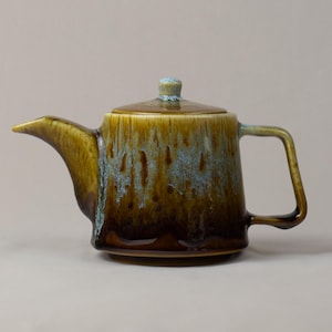 Puede incluir: Una tetera de cerámica con tapa, con un esmalte marrón y azul. La tetera tiene un pico, un asa y un pequeño pomo en la tapa. El esmalte tiene un efecto goteado, creando una textura única. La tetera está diseñada para preparar té.
