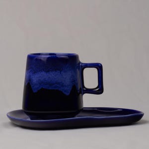Könnte beinhalten: Eine dunkelblaue Keramik-Tasse mit passender Untertasse. Die Tasse hat einen quadratischen Griff und einen Farbverlauf, der von einem helleren Blau oben zu einem tiefen Marineblau unten übergeht. Die Untertasse ist oval geformt.