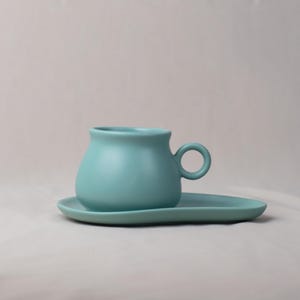 Op de afbeelding: Een turquoise keramische theekop met een afgeronde body en een klein handvat, rustend op een bijpassende ovale schotel. De kop en schotel hebben een matte afwerking. De set is ontworpen voor het serveren van thee of koffie.