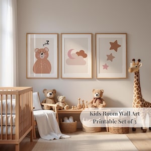 Puede incluir: Tres impresiones enmarcadas de arte mural con un oso, una luna y una nube, y estrellas en tonos rosa y marrón suaves. El texto "Kids Room Wall Art Printable Set of 3" es visible. Una cuna, osos de peluche y una jirafa están en la habitación.