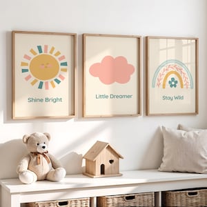 以下が含まれることがあります： 明るいベージュ色の背景に、3つの額入りアートプリント。プリントには、「Shine Bright」の文字が入った笑顔の太陽、「Little Dreamer」のピンクの雲、「Stay Wild」の虹が描かれています。テディベアと木製の家が下にあります。