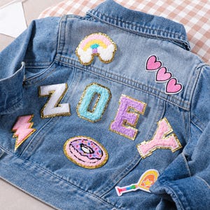 Peut inclure: Veste en jean bleu clair ornée d'écussons colorés. Les écussons comprennent un arc-en-ciel, des cœurs, un éclair, un donut et le nom "ZOEY" épelé avec des lettres individuelles.