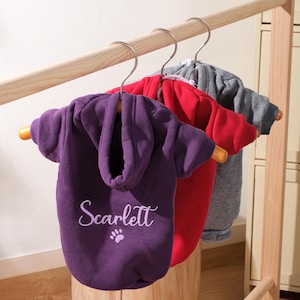Puede incluir: Tres sudaderas con capucha para perros en morado, rojo y gris cuelgan de un perchero de madera. La sudadera morada tiene el nombre "Scarlett" bordado, junto con una huella de pata. Las sudaderas con capucha son de un material suave y tienen capuchas.