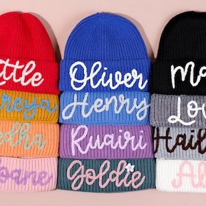 May include: A stack of personalized beanies in various colors, including red, blue, and black. Each beanie features a different name embroidered in a contrasting color. The names include "Little", "Oliver", and "Macey".