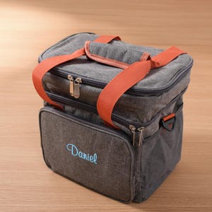 Puede incluir: Bolsa de almuerzo gris aislante con correas naranjas y el nombre "Daniel" bordado en azul. La bolsa tiene dos compartimentos con cremallera, un bolsillo lateral de malla y un asa superior. Diseñada para mantener fríos alimentos y bebidas.