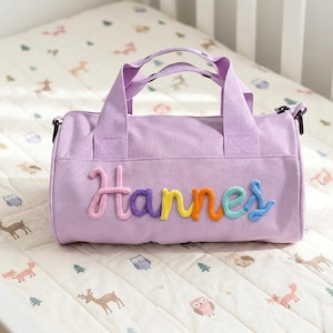 May include: A lilac duffel bag with the name "Hannes" embroidered in rainbow colours. The bag has two handles and a shoulder strap. The bag is on a white patterned surface.