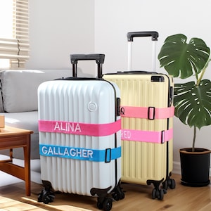 Könnte beinhalten: Zwei Rollkoffer, einer weiß und einer gelb, mit verstellbaren Gepäckgurten. Der weiße Koffer trägt die Namen "ALINA" in Pink und "GALLAGHER" in Blau. Der gelbe Koffer trägt den Namen "RED" in Pink.