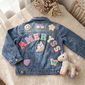 May include: A blue denim jacket featuring colorful embroidered patches including a smiley face, rainbow, flower, unicorn, heart, and butterfly. The name AMERYSS is spelled out in large, multi-colored varsity-style letters across the back.