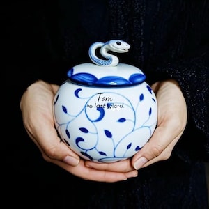 Könnte beinhalten: Eine weiß-blaue Keramikdose mit Deckel, mit einer blauen Schlangenskulptur oben. Die Dose ist mit blauen Blattmustern und dem Text "Tom go last Ward" verziert.