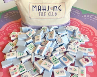 May include: A cream-colored canvas zippered pouch with a lavender base and the text "MAHJONG TILE CLUB" printed on the front. The pouch is surrounded by a large collection of light blue mahjong tiles featuring various colorful graphic illustrations.