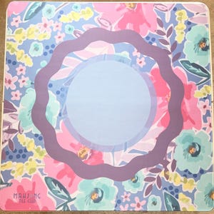 May include: A floral-patterned Mahjong tile mat with a light blue center and a wavy purple border. The mat features pink, blue, and yellow flowers on a light blue background. The words "Mahjong Tile Club" are printed in the lower left corner.