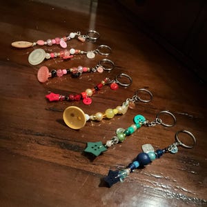 May include: A set of six colorful beaded keychains with different buttons and charms. Each keychain features a variety of beads, including clear, red, green, blue, and yellow beads. The keychains are attached to silver key rings.