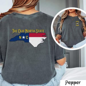 May include: A dark gray t-shirt with a vintage look featuring a graphic of the state of North Carolina with the text "The Old North State" and "N*C" on the back. The front of the shirt has a small graphic of a circle with "N*C" inside.
