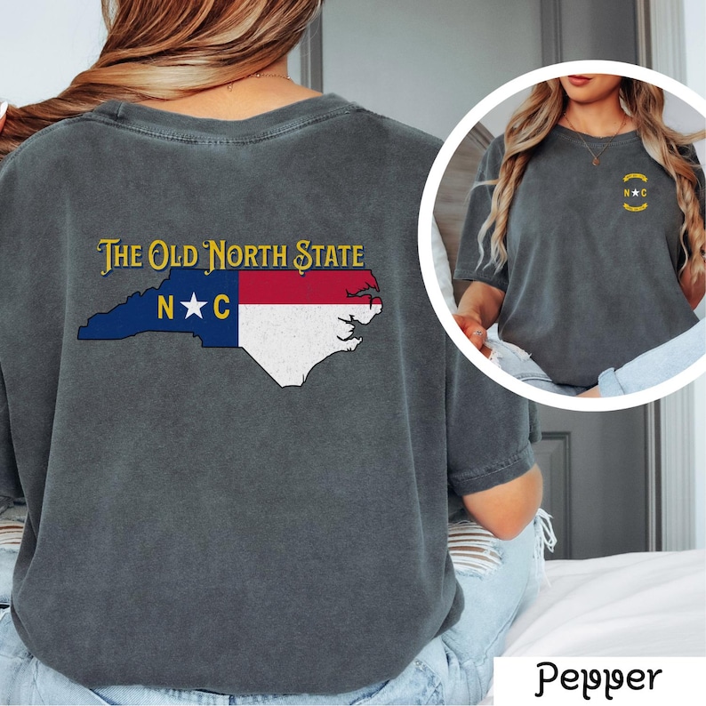May include: A dark gray t-shirt with a vintage look featuring a graphic of the state of North Carolina with the text "The Old North State" and "N*C" on the back. The front of the shirt has a small graphic of a circle with "N*C" inside.