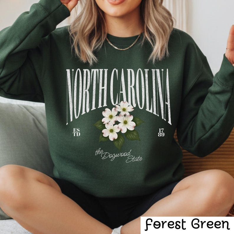 May include: Forest green sweatshirt with the text "NORTH CAROLINA" in a vintage style, featuring white dogwood flowers. The sweatshirt also includes the text "ESTD" and "1789" with "the Dogwood State" below the flowers.