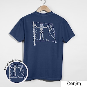 May include: Denim blue t-shirt with a white graphic of a skeleton on a flag. The flag design is on the back of the shirt. A smaller version of the graphic is on the front left chest. The word "Denim" is at the bottom right.